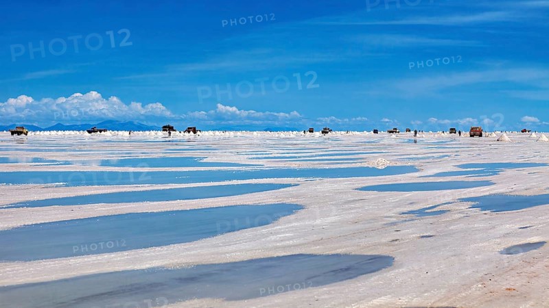 Puddles on a salt flat with vehicles on the horizon and clouds in the sky, The endless expanses of the salt plain of the Salar de Uyuni in Bolivia