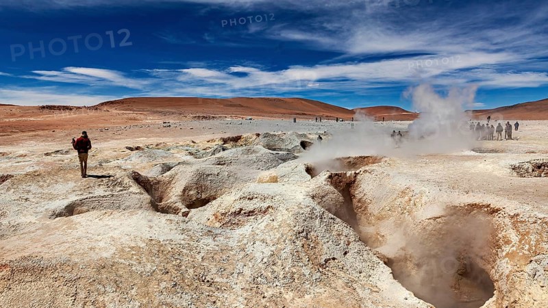 Lone hiker in a desert landscape with a smoking geyser and blue sky, The mud volcanoes and fumaroles on the Altiplano in Bolivia