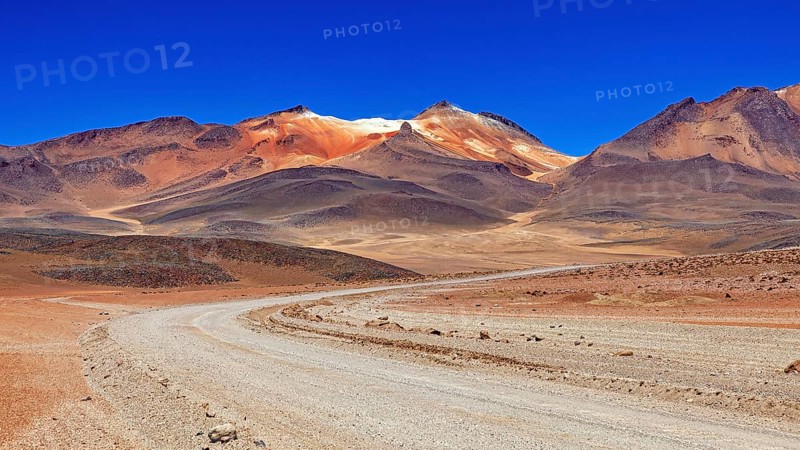 A winding sandy road through a dry landscape with reddish mountain peaks and blue skies, The landscape of the Altiplano in Bolivia