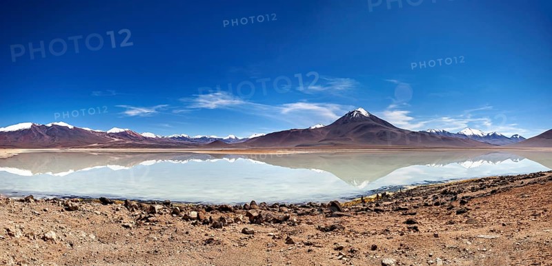 Wide view of a volcano with a reflecting lake and rocky shore under a blue sky, The Laguna Colorada in Bolivia