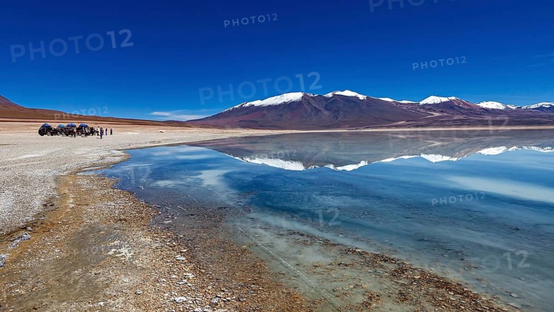 Panoramic view of a lake with snow-capped peaks, people and vehicles on the shore, The Laguna Colorada in Bolivia