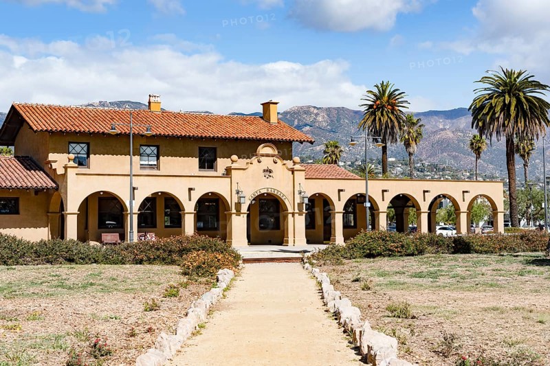 The historic Santa Barbara train station, featuring Spanish Colonial Revival architecture with a red tile roof and arches, set against scenic mountains and palm trees, California, USA