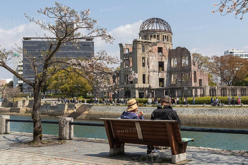 Tourists sitting on a bench under blossoming cherry trees, Sakura, Atomic Bomb Dome behind them, ruins of the former Chamber of Commerce and Industry building, landmark, UNESCO World Heritage Site, peace memorial and memorial to the victims of the atomic bombing of Hiroshima in the Peace Park on the Otagawa River, Naka Ward, Hiroshima, Japan
