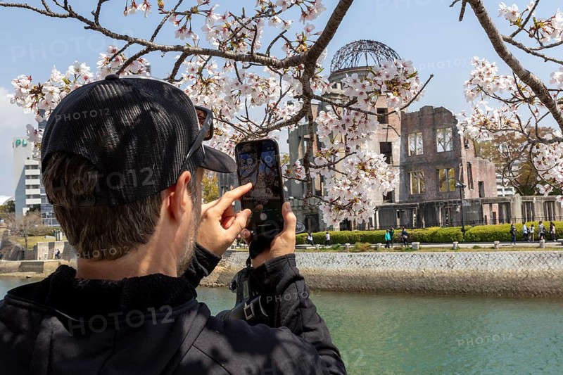 Tourist taking a photo of cherry blossoms with a smartphone, sakura, behind it atomic bomb dome, ruins of the former building of the Chamber of Commerce and Industry, landmark, UNESCO World Heritage Site, peace monument and memorial to the victims of the atomic bombing of Hiroshima in the Peace Park on the Otagawa River, Naka Ward, Hiroshima, Japan