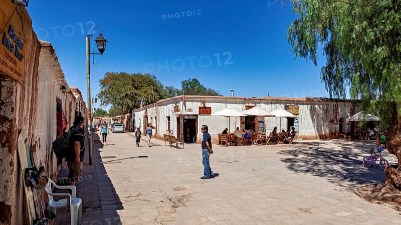 People sit under umbrellas on a lively street next to typical local buildings in the sunshine, The town of San Pedro de Atacama in Chile