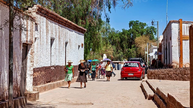 Pedestrians in summer clothes walk along a street with white buildings and a red car, The town of San Pedro de Atacama in Chile