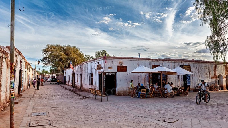 People move along a street with old buildings under partly cloudy skies and cyclists, The town of San Pedro de Atacama in Chile