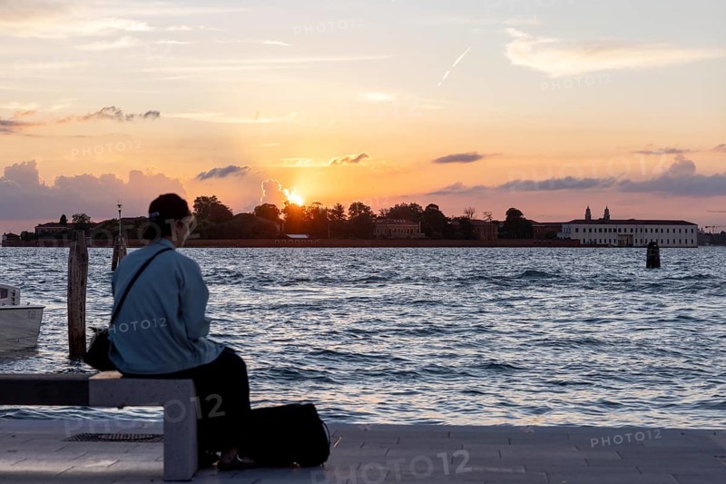 Venice, Italy - 2 September 2025: Passengers sit on the jetty at the Lido at sunset, waiting for the arrival of a boat to take them to another island, while a film poster is seen in the foreground, during the 82nd Venice International Film Festival