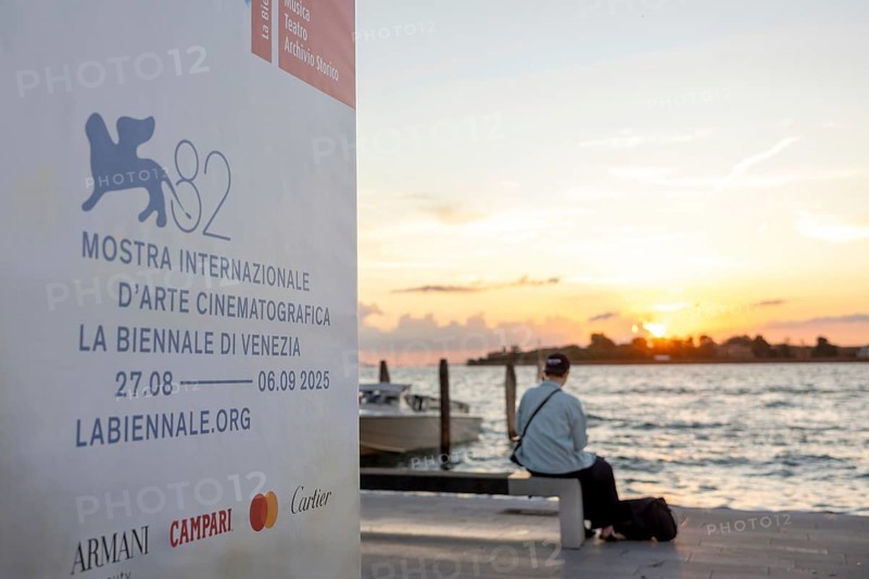 Venice, Italy - 2 September 2025: Passengers sit on the jetty at the Lido at sunset, waiting for the arrival of a boat to take them to another island, while a film poster is seen in the foreground, during the 82nd Venice International Film Festival