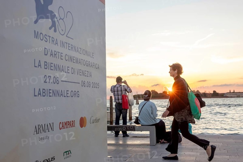 Venice, Italy - 2 September 2025: Passengers sit on the jetty at the Lido at sunset, waiting for the arrival of a boat to take them to another island, while a film poster is seen in the foreground, during the 82nd Venice International Film Festival