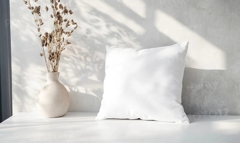 A white pillow sits on a table next to a vase of flowers. The pillow is unmade and the flowers are wilted. Scene is somewhat sad and lonely. AI generated
