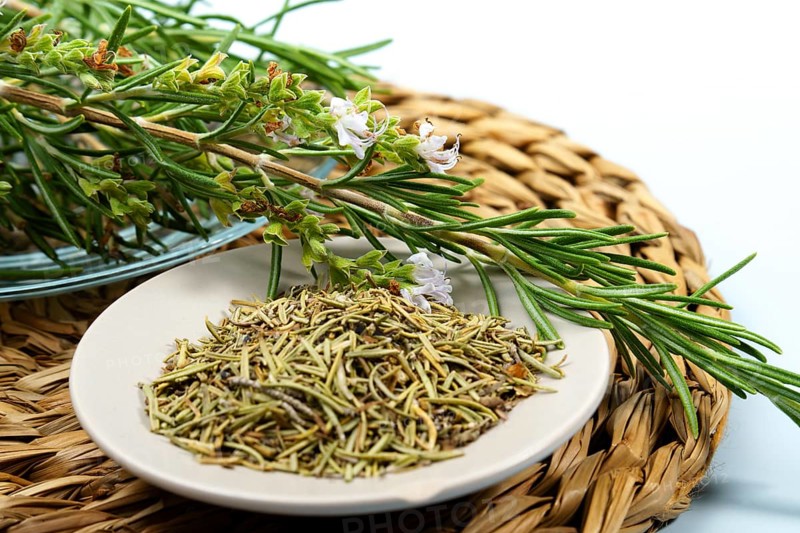 Fresh rosemary sprigs with flowers lie on a plate of dried rosemary, ready for culinary or medicinal use