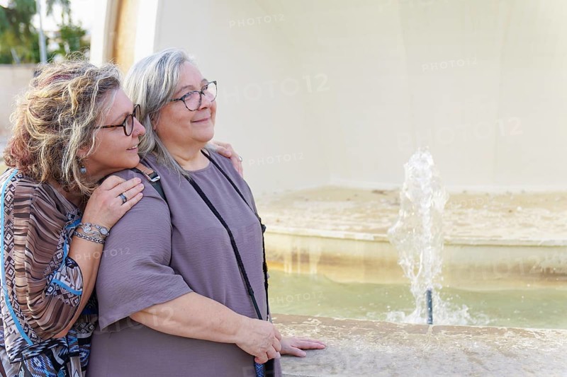 Two happy senior women hugging each other near a fountain in a city park, enjoying a sunny day together