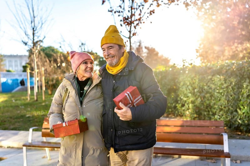 Elderly couple holding christmas presents smiling and walking in a city park during a sunny winter day