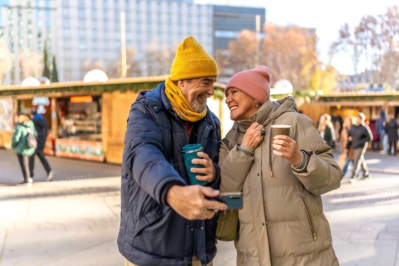 Smiling senior couple taking a selfie and drinking hot beverages at a christmas market in the city