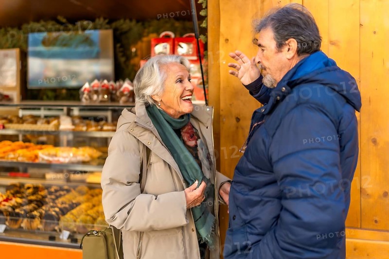 Cheerful elderly couple enjoying christmas time together, buying sweets and pastries at a wooden stall in a traditional christmas market