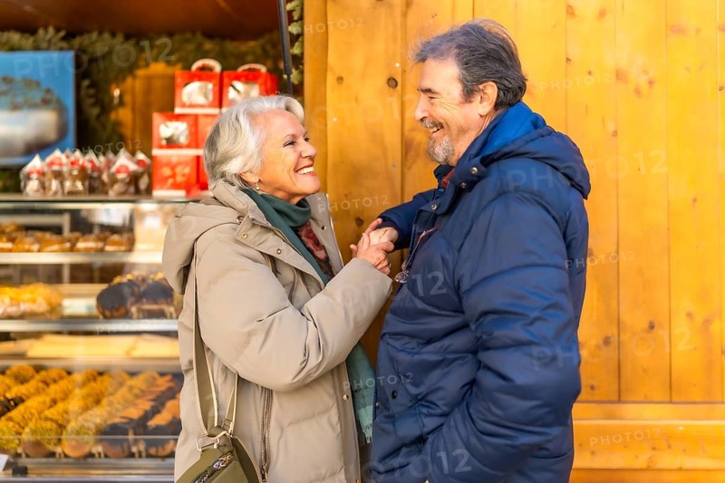 Smiling senior couple holding hands and looking at each other in front of a christmas bakery shop, enjoying winter holidays together