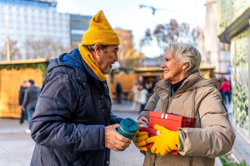 Happy senior couple exchanging christmas gifts and enjoying a warm beverage at a bustling outdoor christmas market in the city