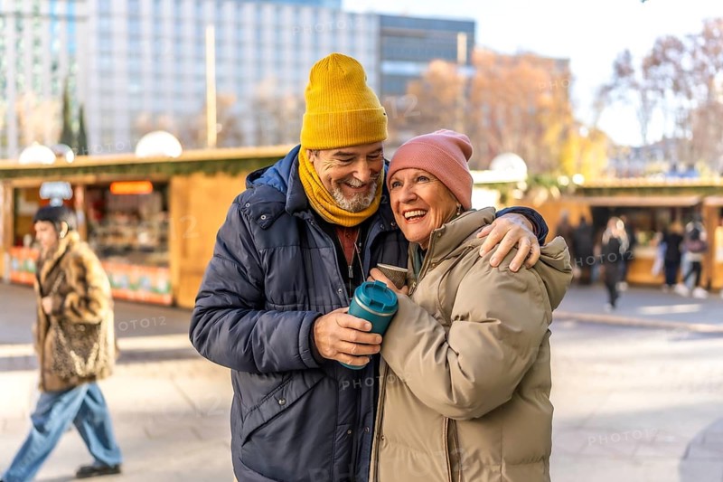 Elderly couple sharing warm drinks while enjoying the festive atmosphere of an outdoor christmas market, embracing and smiling joyfully