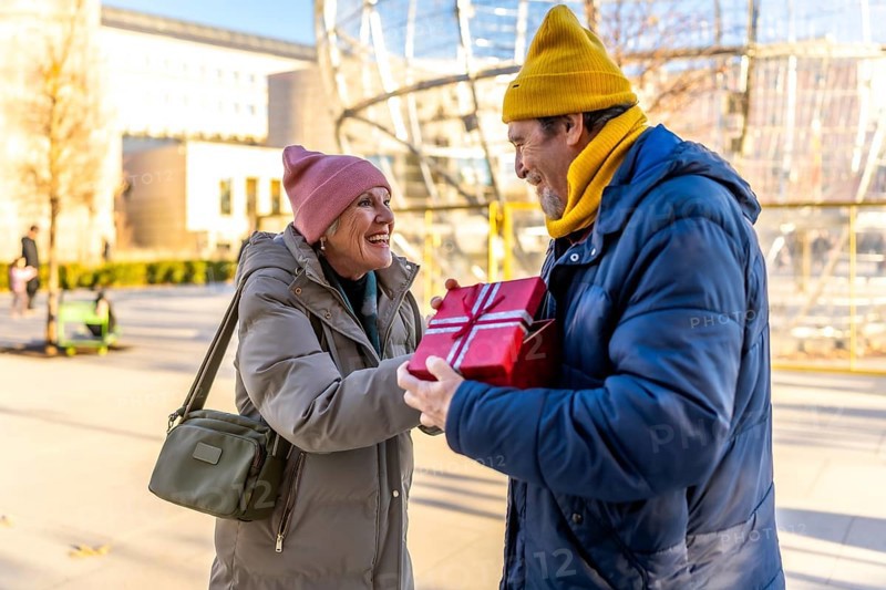 Elderly man joyfully giving a christmas present to a smiling woman in a sunny winter city park, celebrating love and togetherness