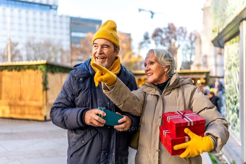 Cheerful senior couple holding christmas gifts and pointing at something interesting at the christmas market in the city