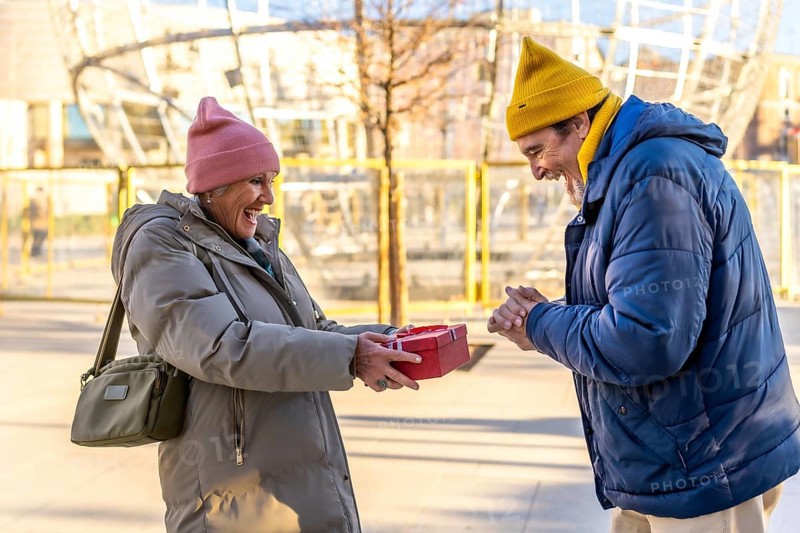 Cheerful senior woman giving a christmas present to her excited husband in a city during wintertime