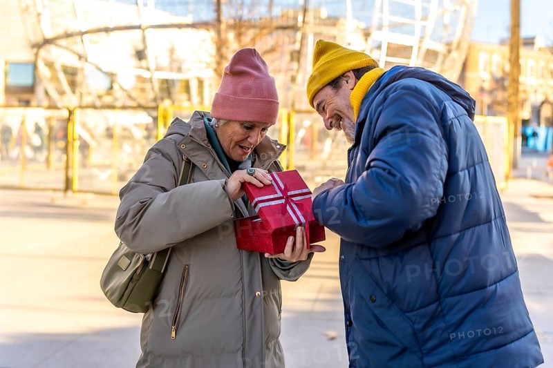 Happy senior couple sharing a joyful moment while opening a festive christmas gift box in a charming winter city setting