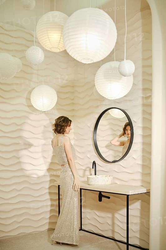 A woman dressed in a sparkling gown admires her reflection in a round mirror. Soft, textured walls and pendant lights create a serene atmosphere in a modern bathroom