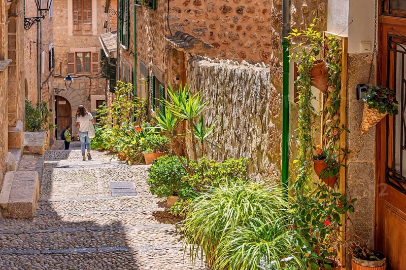 Alone woman walking in an idyllic cobblestone alley in an old village with green plants and flowers in pots outside a door on a sunny summer day, Fornlalutx, Mallorca, Spain