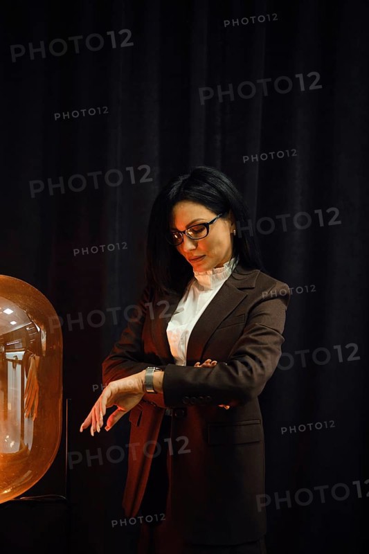 A woman dressed in a fashionable brown suit stands near a glowing lamp. She checks her watch with a contemplative expression, set against a dark backdrop that contrasts with the warm light