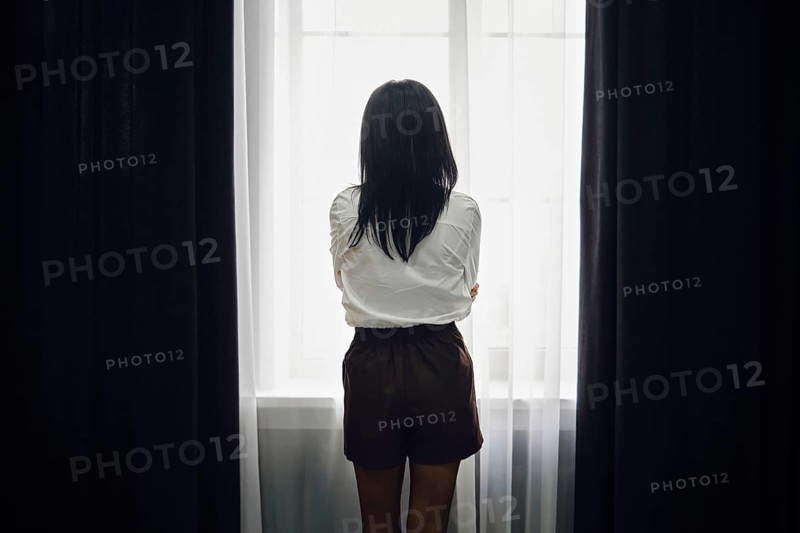 A woman stands with her back to the viewer, gazing out of a bright window. The soft afternoon light filters through sheer curtains, creating a tranquil atmosphere in the room