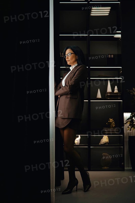 A confident woman in brown jacket, mini skirt and glasses stands with her arms crossed in an office