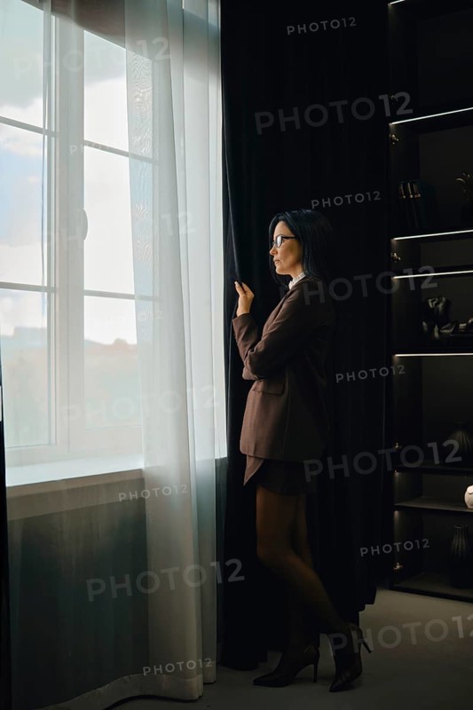 A woman dressed in a tailored suit stands by a large window, pulling back sheer curtains. She appears thoughtful as she gazes outside, surrounded by elegant, dark furnishings