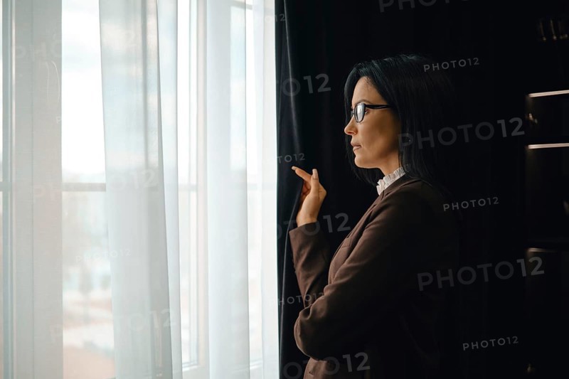 A businesswoman stands by a window in a modern office, gazing thoughtfully outside. She wears glasses and a stylish outfit, with soft natural light illuminating the room