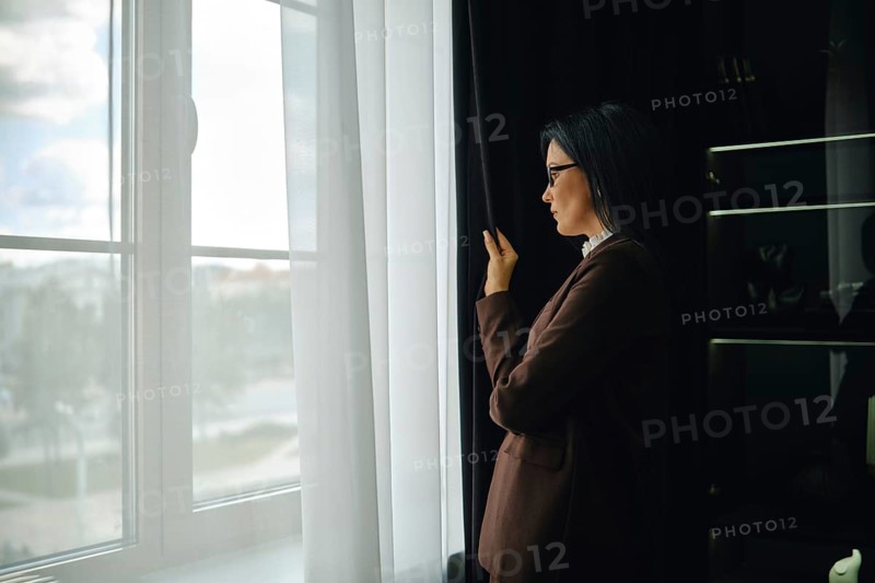 Woman in brown jacket gazes out window while reflecting on the day in a modern setting