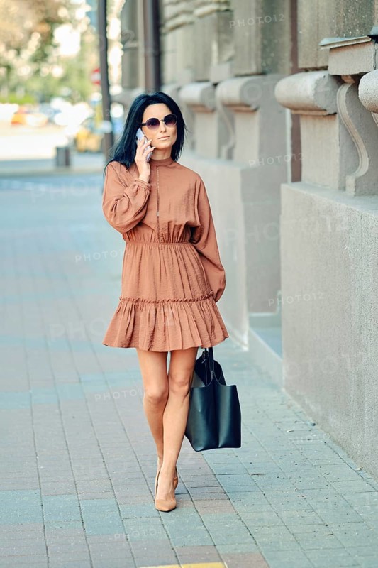 A fashionable woman strolls down the city sidewalk wearing a brown dress and sunglasses, carrying a black handbag. Warm sunlight illuminates the bustling urban surroundings