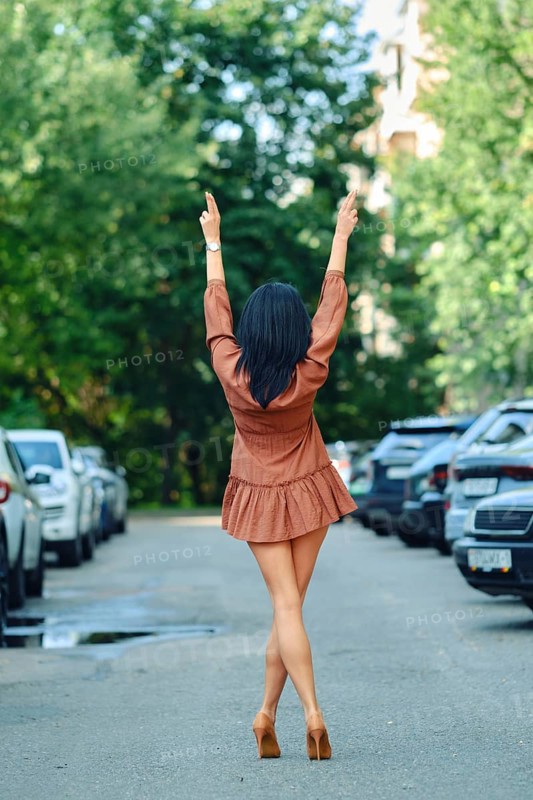 Rear view of a woman walks down a tree-lined street, wearing a fashionable brown outfit and high heels. Her arms are raised in a joyful gesture as she enjoys the sunny day in the urban setting