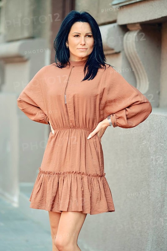 A woman stands with her arms on waist on a city sidewalk, wearing a chic, loose-fitting dress. Her long dark hair frames her face as she strikes a poised pose against a minimalist backdrop