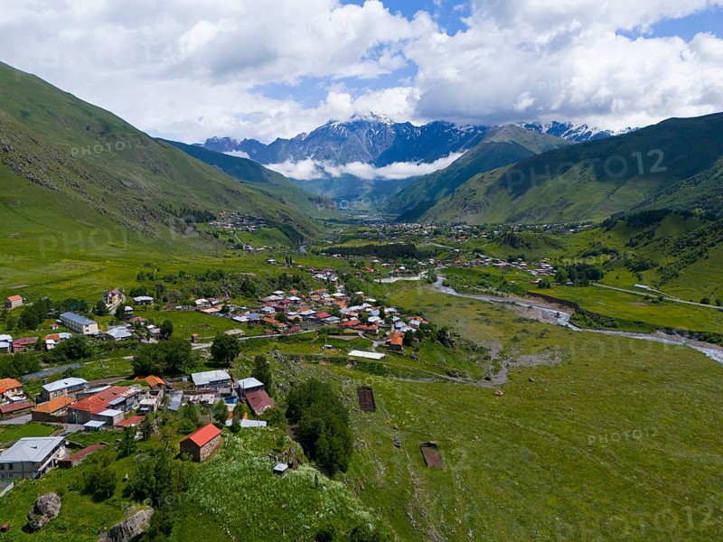 Village in a wide valley between green mountains with clouds in the sky, landscape near Kazbegi, Qasbegi, Stepantsminda, Georgian Military Road, Mtskheta-Mtianeti province, Mount Shani in the background, located in Ingushetia, Russia, High Caucasus, Georgia