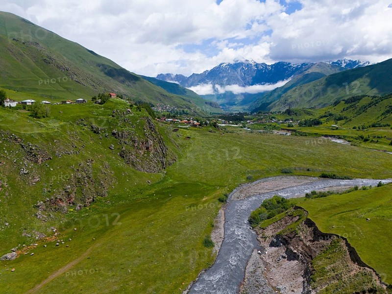 River flowing through green valley, surrounded by mountains and clouds, near a small village, aerial view, Terek River, Kazbegi, Qasbegi, Stepantsminda, Georgian Military Road, Mtskheta-Mtianeti Province, Shani Mountain in the background, located in Ingushetia, Russia, High Caucasus, Georgia