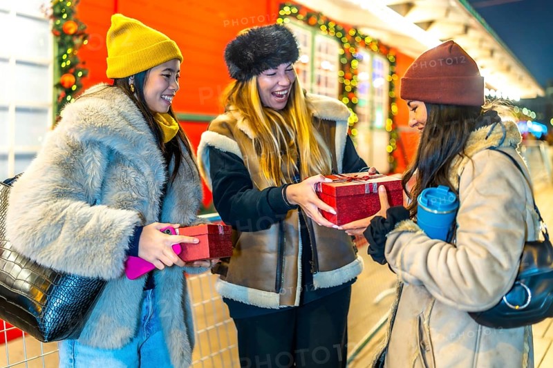 Three joyful women exchanging christmas gifts while enjoying the festive atmosphere of a beautifully decorated night market