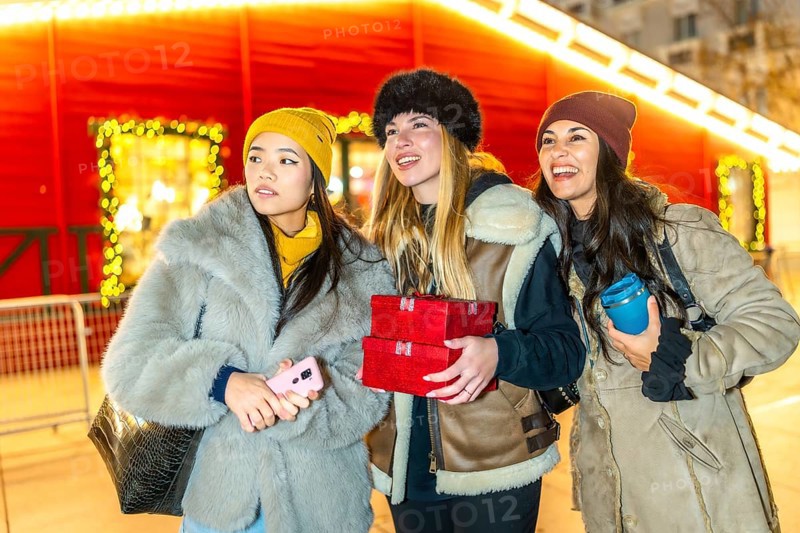 Three happy female friends enjoying christmas night market in the city, holding gifts and a reusable mug
