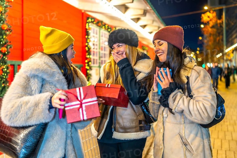 Three cheerful women exchanging christmas gifts and enjoying hot beverages at a vibrant night market, celebrating the festive season together