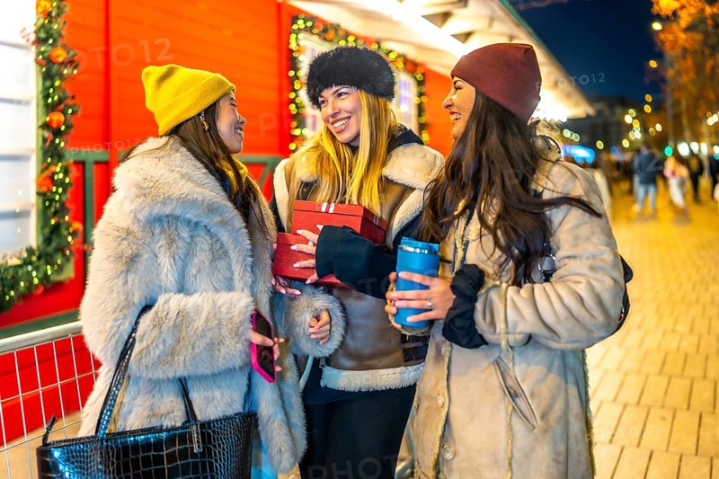 Three cheerful women holding christmas gifts and hot drinks, enjoying christmas shopping at a decorated night market