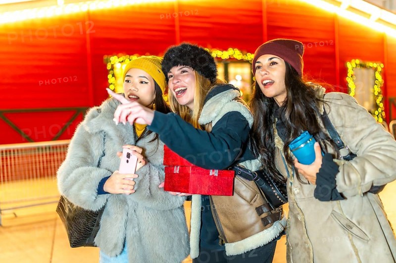 Three young women holding christmas gifts and a smartphone are enjoying the christmas spirit, pointing at something interesting in a brightly lit market stall