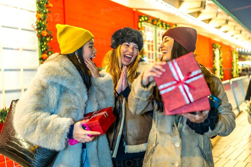 Three cheerful women exchanging christmas presents at a decorated outdoor night market, enjoying the festive atmosphere