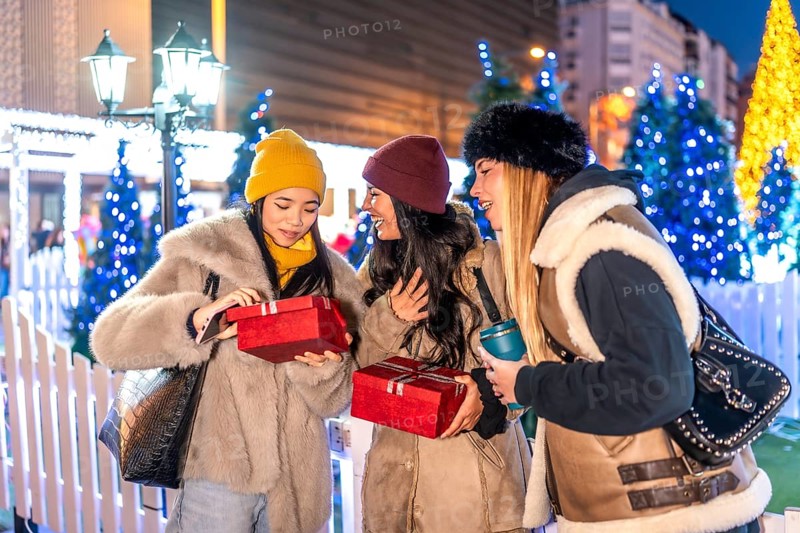 Cheerful young women exchanging christmas gifts at a decorated christmas market in the city at night