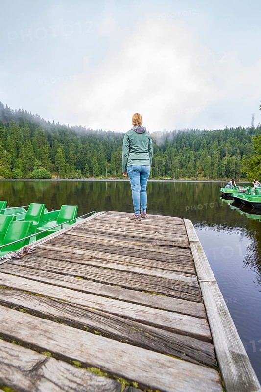 Woman standing alone on a wooden jetty in front of a quiet lake surrounded by forest, Mummelsee, Seebach, Ortenau District, Black Forest, Germany