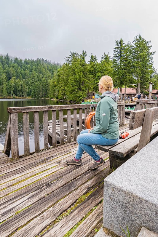 A woman sits relaxed on a wooden bench overlooking the tranquil lake and forest, Mummelsee, Seebach, Ortenau District, Black Forest, Germany