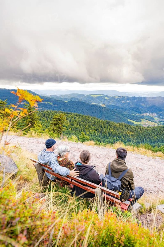 Three people on a bench enjoy the wide view over wooded mountains and valleys, Mummelsee, Seebach, Ortenau district, Black Forest, Germany